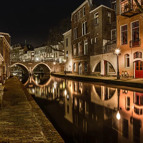Sfeervol nachtbeeld van een gracht in een historische binnenstad met brug, verlichte gevels en spiegeling in het water.