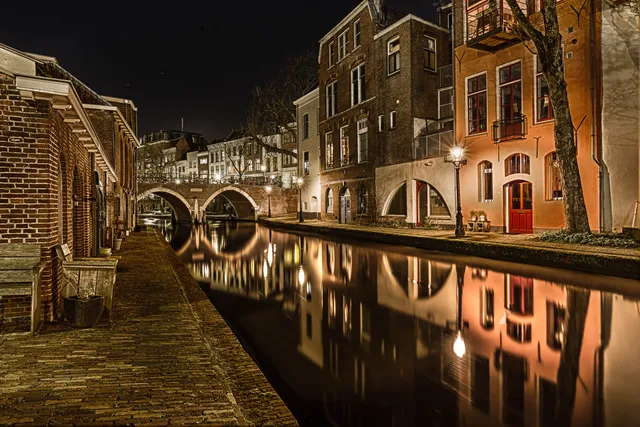 Sfeervol nachtbeeld van een gracht in een historische binnenstad met brug, verlichte gevels en spiegeling in het water.