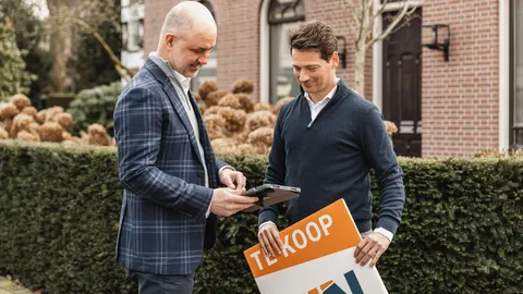 Twee makelaars met een tablet en een te-koopbord bij een woning