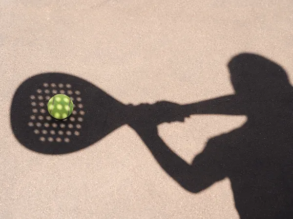 Schaduw van een padelspeler met racket en een groene bal op een zonnige ondergrond.